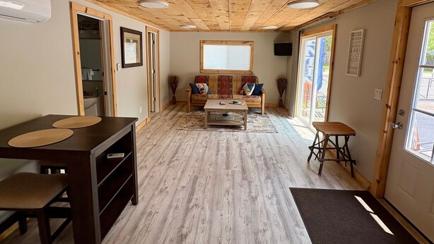 A room with a wooden ceiling, a sofa, a coffee table, a small table with chairs, and a door leading outside. - Charming 2-bedroom cabin with WiFi, AC in serene Oscoda (Oscoda)