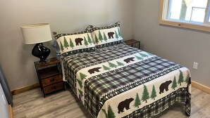 A bedroom with a bed featuring a bear and tree pattern, a bedside table with a lamp, and a window with a view of the outside. - Charming 2-bedroom cabin with WiFi, AC in serene Oscoda (Oscoda)