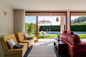 A modern living room with a sofa, a red armchair, and a dining table. There is a large sliding glass door leading to a pool area with outdoor furniture.