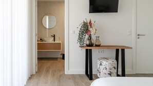 A modern hotel room with a wooden floor, a round mirror, a flat-screen TV, a wooden table with a vase of flowers, and a floral-patterned ottoman.