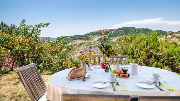 An outdoor dining table set with a tablecloth, plates, cups, and a loaf of bread, overlooking a scenic landscape.