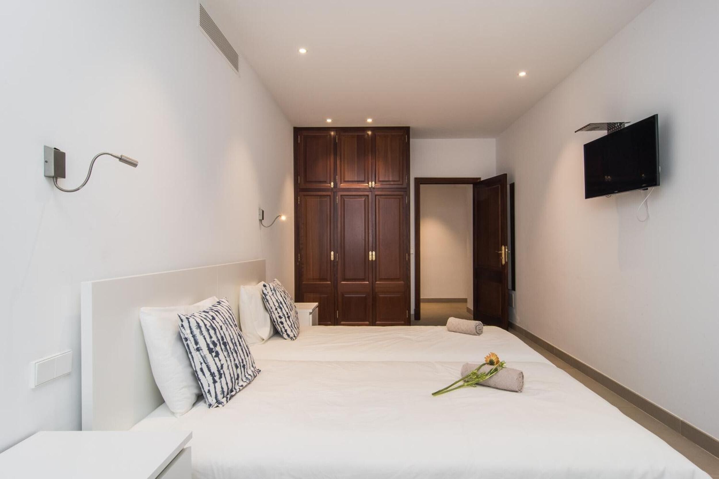 A modern hotel room with a large bed, a wardrobe, a TV mounted on the wall, and a small bedside table.