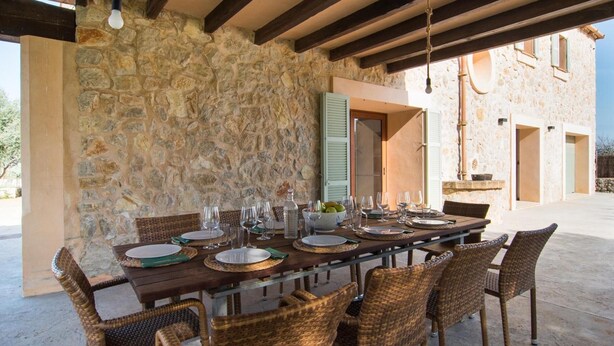 Un área de comedor al aire libre con paredes de piedra, sillas de mimbre y una larga mesa de madera lista para una comida.