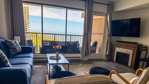 Ein Wohnzimmer mit Sofa, Sessel, Couchtisch und Kamin. Ein großes Fenster bietet Blick auf den Ozean und einen Balkon.