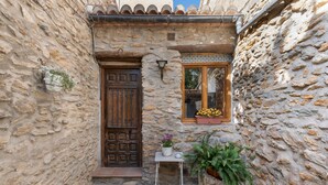 Un edificio de piedra con una puerta de madera, una ventana pequeña con una planta en maceta, y una mesa de metal también con una planta en maceta.