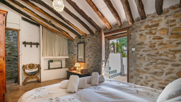 Un dormitorio con pared de piedra, vigas de madera, cama con ropa blanca, mesita de noche con lámpara y ventana con cortinas.