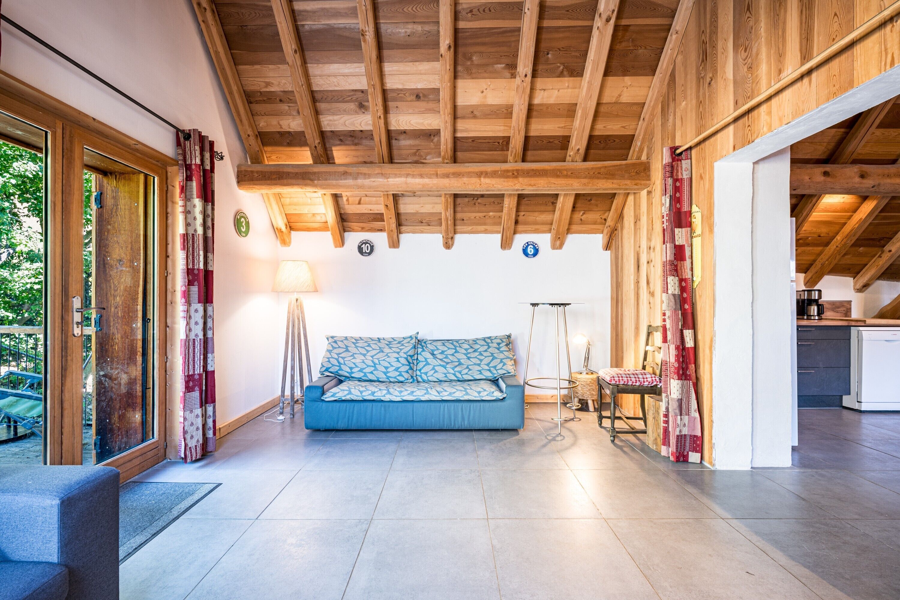 A wooden ceiling with exposed beams, a blue sofa with patterned cushions, a chair, and a small table with a lamp.