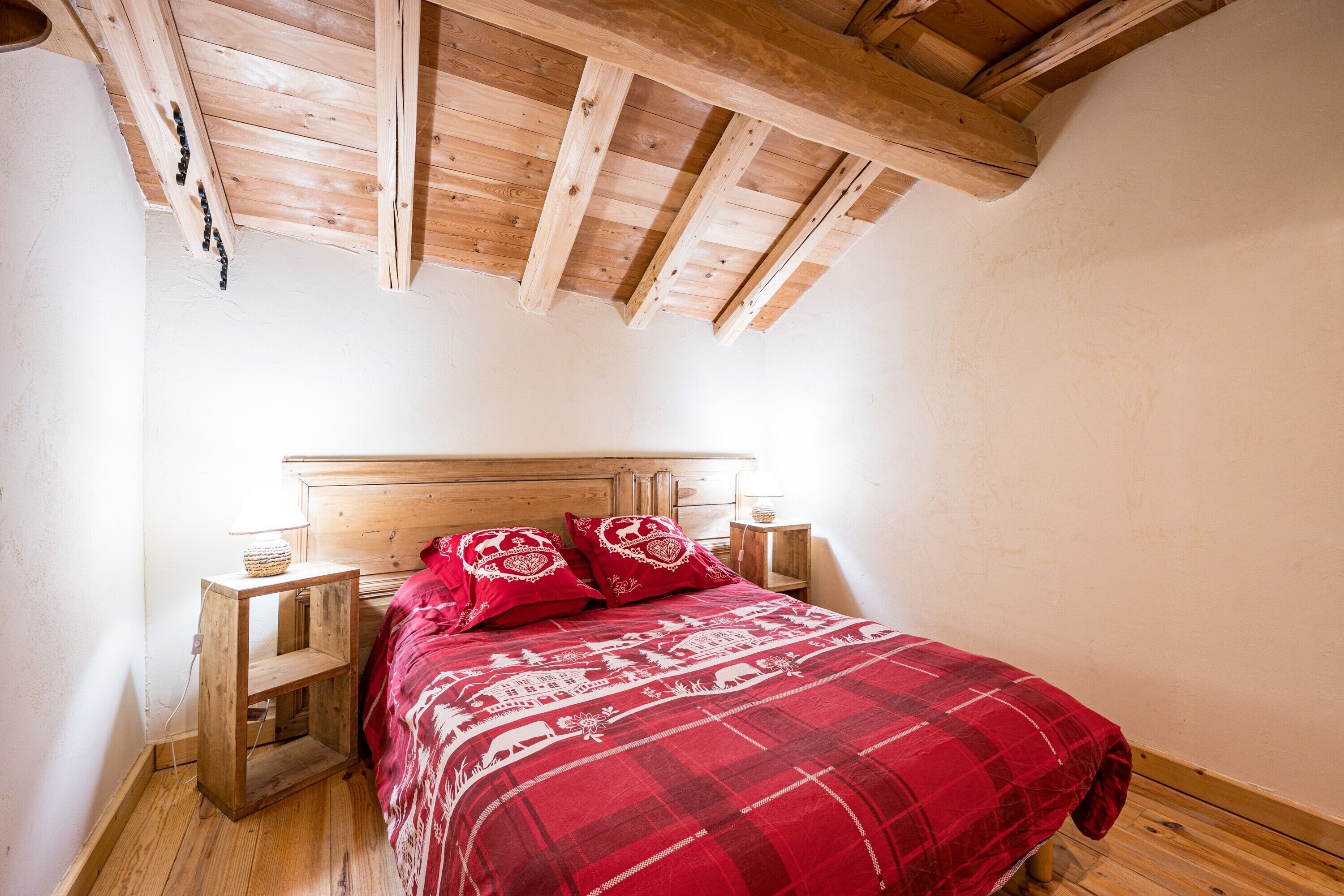 A bedroom with a wooden headboard, a red plaid bedspread, two bedside tables with lamps, and a wooden floor.