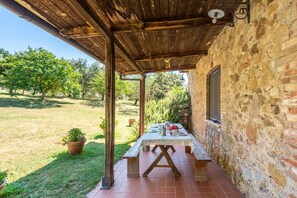 An outdoor patio with a wooden table and benches, surrounded by a stone wall and a grassy area with trees.