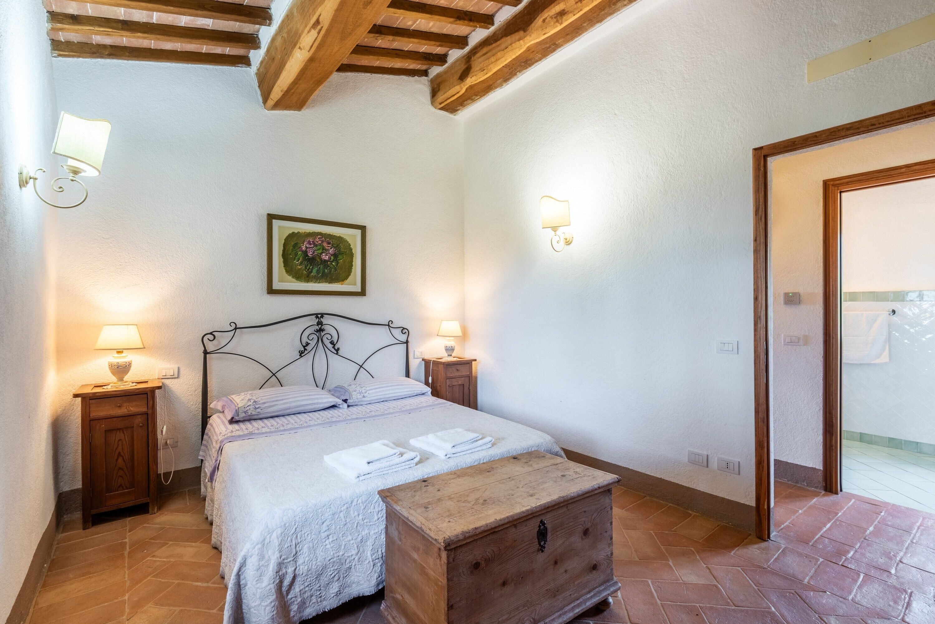 Une chambre à coucher avec un lit, des lampes de chevet, une commode en bois, un tableau au mur et une porte sur le côté droit.