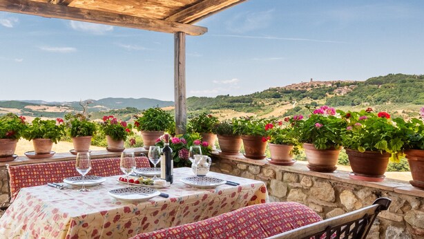 Un área de comedor al aire libre con una mesa dispuesta para dos, rodeada de plantas en macetas y con vistas a una ladera panorámica.