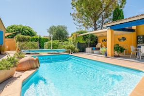 A swimming pool with a sunshade, surrounded by potted plants and a patio area with outdoor furniture.