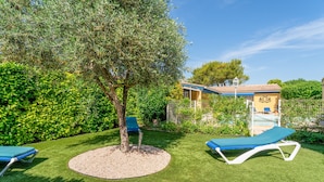 A well-maintained garden with artificial grass, two lounge chairs, a tree, and a building with a blue roof in the background. - Holiday Home “L'Olivier” with Shared Pool, Wi-Fi & Air Conditioning (L'Isle-sur-la-Sorgue)