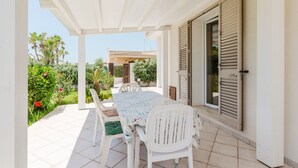 A covered patio with a table and chairs, a door with shutters, and a garden with various plants and flowers.