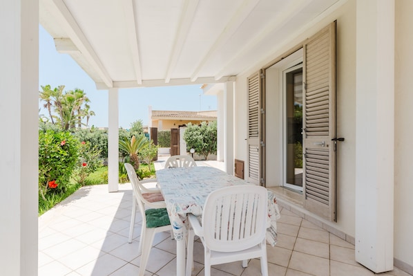 A covered patio with a table and chairs, a door with shutters, and a garden with various plants and flowers.