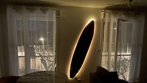 A dimly lit room with a round table and chairs, a surfboard-shaped light fixture, and a view through a window with sheer curtains. - Modern studio - terrace - pool (Villeneuve-Loubet)