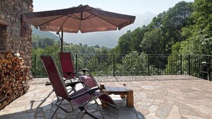 A stone patio with a wooden table and two red lounge chairs, an umbrella, and a stack of firewood. - Deluxe Room "Chalet Cuore Selvatico" with Mountain View, Terrace, and Private Garden (Monno)
