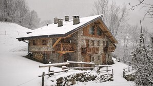 Une cabane rustique en pierre, dotée de volets en bois, située dans une région montagneuse enneigée.