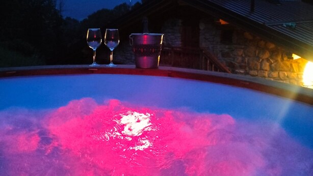 Un bain à remous avec deux verres et une carafe, un bâtiment en pierre et une chaîne de montagnes en arrière-plan.