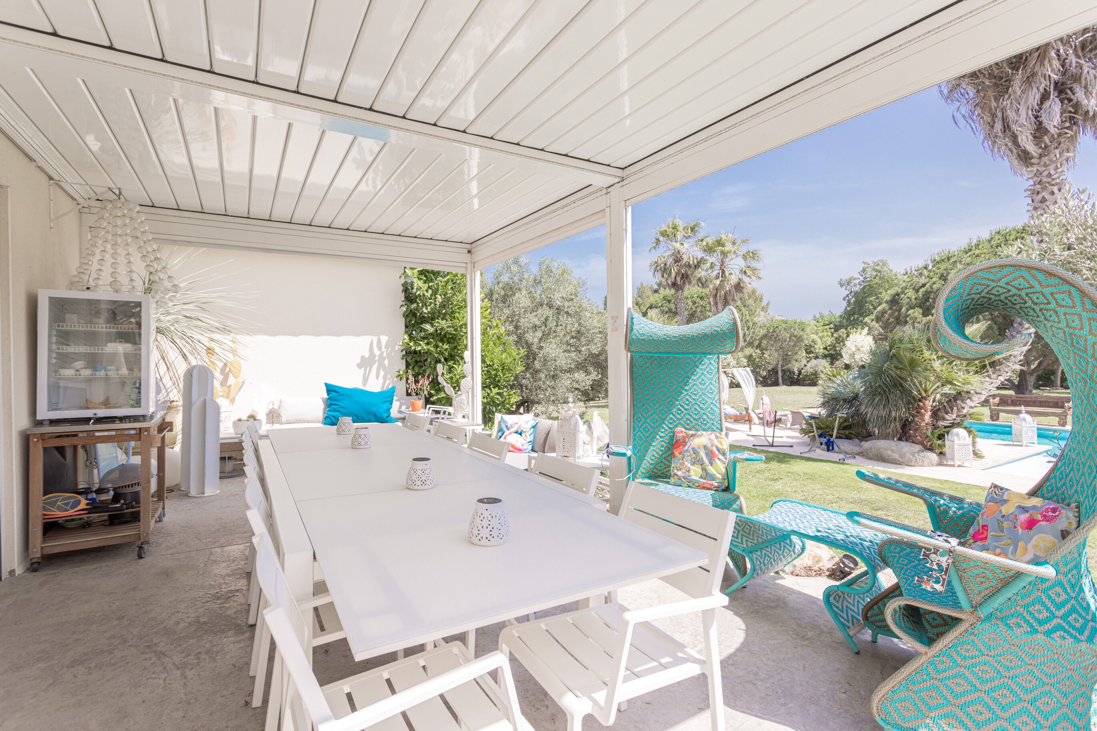 An outdoor patio with a white table and chairs, a TV on a cart, and a view of a pool and garden.