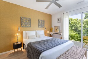 Une chambre avec un lit, un ventilateur de plafond, une table de chevet, une lampe, des œuvres d’art au mur et une vue sur la verdure à l’extérieur.