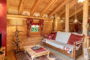 Interior de una cabaña de madera con un sofá, una mesa con un cuenco, una lámpara y una ventana con cortinas.