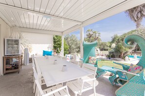 Une terrasse extérieure avec une table et des chaises blanches, un téléviseur posé sur un chariot, et une vue sur une piscine et un jardin.