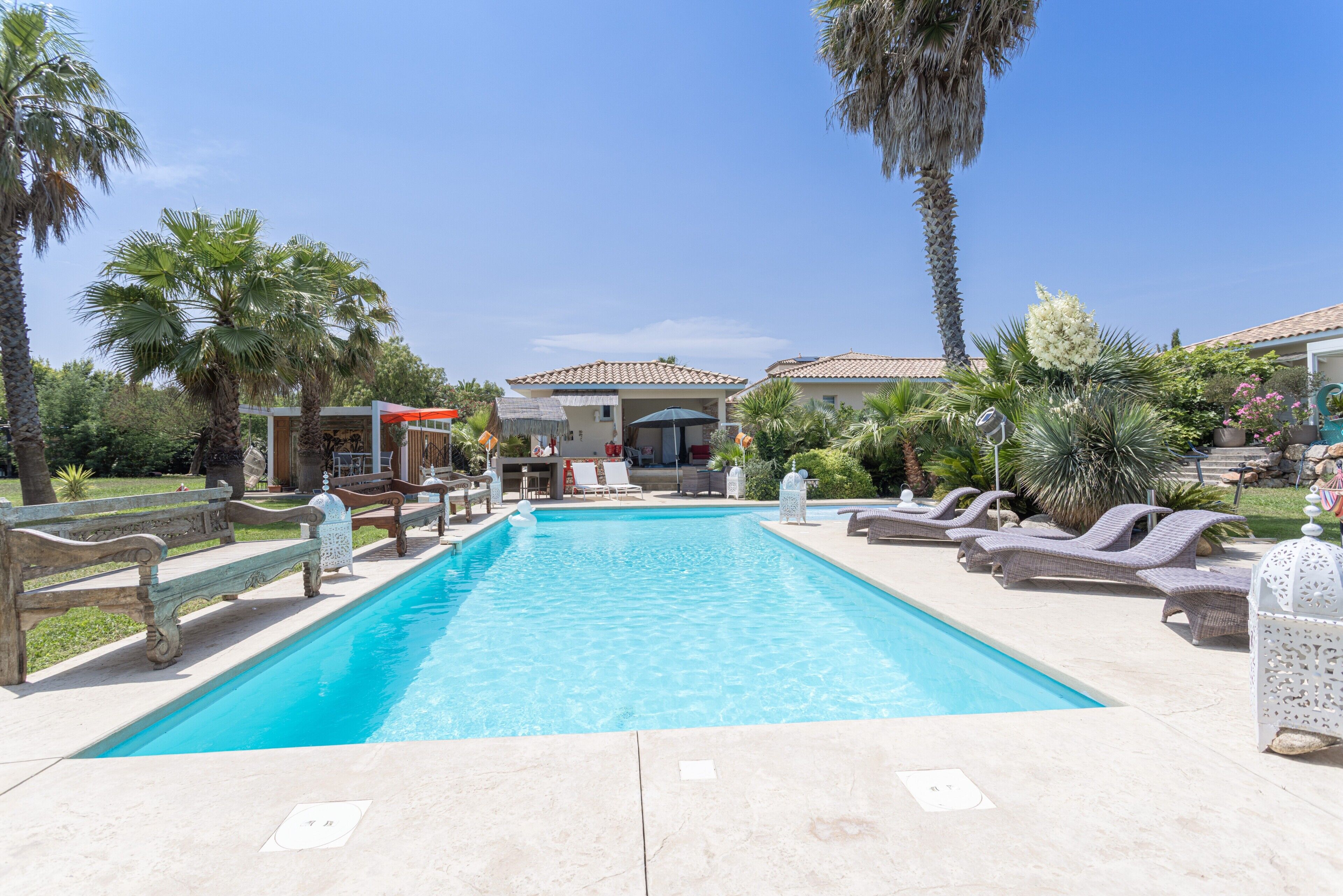 A swimming pool with lounge chairs, palm trees, and a building with a tiled roof in the background.