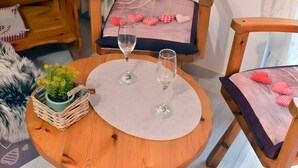 Een ronde houten tafel met een witte placemat, twee wijnglazen, een kleine potplant en een stoel met een kussen met hartvormige kussentjes.