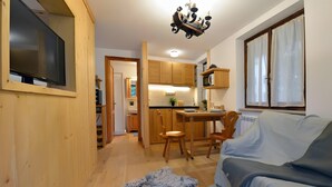 Um quarto aconchegante com piso de madeira, um sofá, uma mesa de jantar com cadeiras, uma TV fixada em uma parede de madeira e uma cozinha com armários de madeira e micro-ondas.