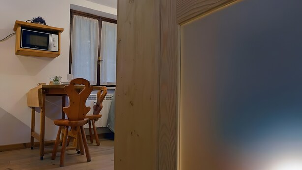 Uma mesa de jantar de madeira com cadeiras, um micro-ondas numa prateleira e uma janela com cortinas.