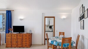 A dining area with a table, chairs, a television on a wooden cabinet, and a kitchen in the background. - Colmeia AL - Casa Buzz: Apartamento com Vista Mar em Armação de Pera (Unknown)