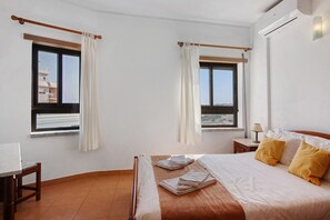 A hotel room with a bed, a desk, two windows with curtains, a nightstand, and an air conditioning unit. - Colmeia AL - Casa Pólen (Armação de Pêra)