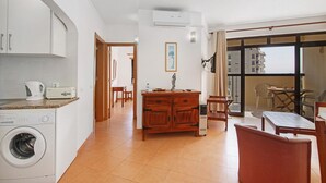 A modern kitchen with a washing machine, a dining area with a table and chairs, and a view through a large window.