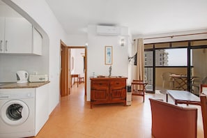 A modern kitchen with a washing machine, a dining area with a table and chairs, and a view through a large window. - Colmeia AL - Casa Pólen (Armação de Pêra)