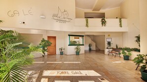 A spacious indoor area with a tiled floor, potted plants, a bench, and a wall with a ship drawing and the text 'GALÉ'.