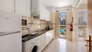 A modern kitchen with a washing machine, a refrigerator, and a window overlooking a pool. - Colmeia AL - Casa Néctar (Armação de Pêra)