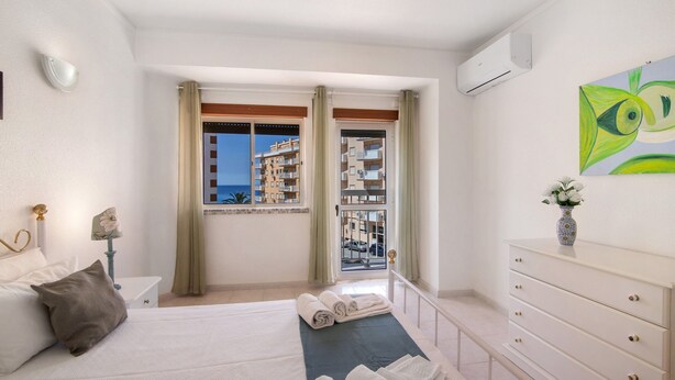 A hotel room with a bed, a dresser, a window with curtains, and a view of buildings. - Colmeia AL - Casa Néctar (Armação de Pêra)