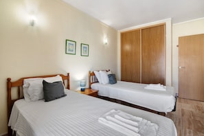 Dos camas individuales con cabeceros de madera, ropa de cama blanca y un armario de madera en una habitación con paredes color beige y suelo de baldosas.