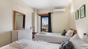 A hotel room with two beds, a dresser, a mirror, an air conditioner, and a view of buildings. - Colmeia AL - Casa Enxame (Armação de Pêra)