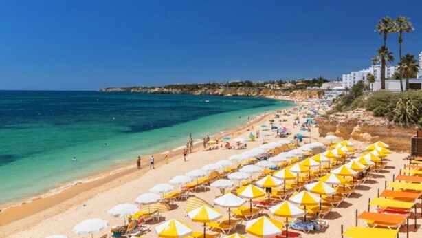 A beach with rows of sun umbrellas, clear blue water, and buildings in the background. - Colmeia AL - Casa Enxame (Armação de Pêra)