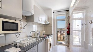 A modern kitchen with a built-in microwave, kettle, and washing machine, a black countertop, and a view of the outside through a glass door. - Colmeia AL - Casa Mel: Vista Mar e Conforto em Armação de Pera (Unknown)