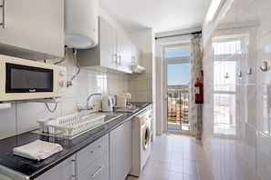 A modern kitchen with a built-in microwave, kettle, and washing machine, a black countertop, and a view of the outside through a glass door.