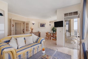 A living room with a sofa, a coffee table, a dining area with chairs, a television, and a glass door leading outside.