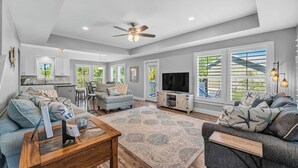 A spacious living room with a large sofa, a coffee table, and a TV. There is a dining area with a table and chairs, and a kitchen in the background. - 4 Bedroom Pet Friendly House in Fort Morgan - Free Daily Activities! (Gulf Shores)