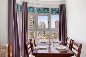 A dining table set for four with a view of buildings and the sea through a bay window. - Colmeia AL - Casa Cera: Apartamento com Vista Mar em Armação de Pera (Armação de Pêra)