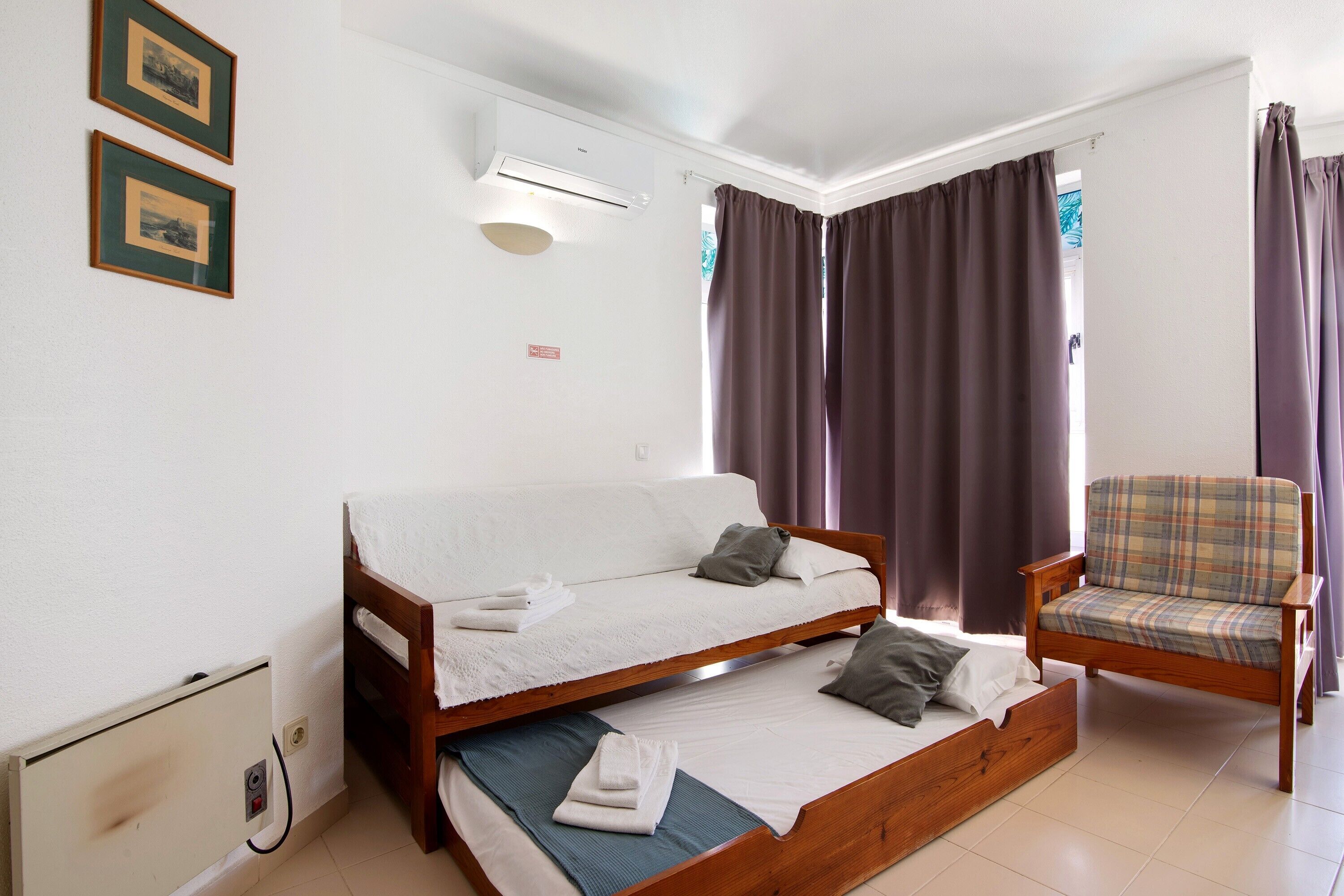 A room with a bed, a chair, an air conditioning unit, and curtains.