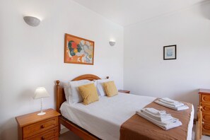 Una cama bien hecha con una colcha blanca y marrón, dos almohadas, una mesita de noche con lámpara y un aparador de madera.