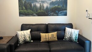 Un canapé en cuir noir agrémenté de trois coussins décoratifs, un grand tableau représentant un paysage de forêt et de montagnes accroché au mur, et un thermostat fixé au mur, à droite.
