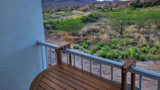 Une terrasse en bois, dotée d’une balustrade blanche, offre une vue sur un paysage désertique parsemé d’arbustes verts et dominé par des montagnes lointaines.